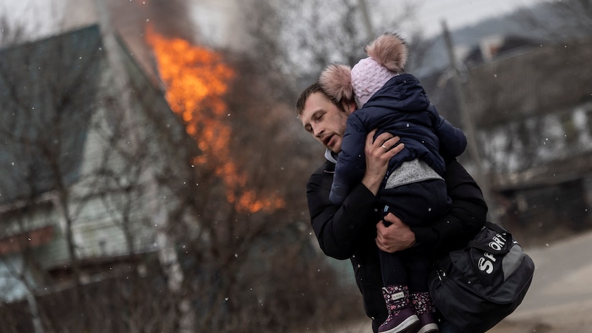 A man runs with a child in his arms, away from a building on fire.