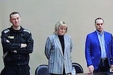 A man dressed in all black with his arms crossed, a woman wearing a blazer and a man wearing a blue suit stand behind a table.