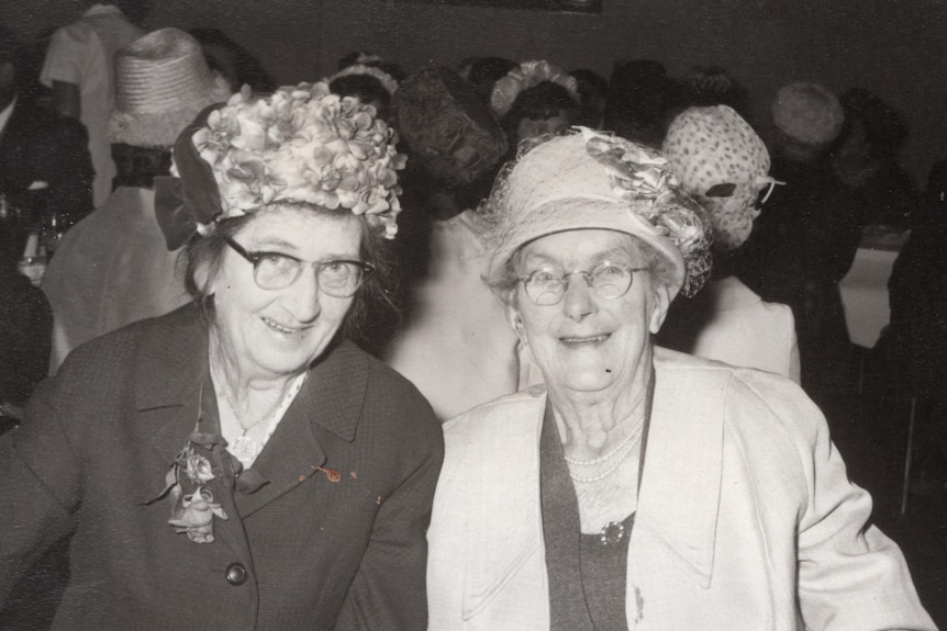 two ladies with hats on sit together smiling at the camera in a black and white photo