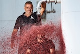 Two middle-aged men - one is pouring red wine from a tank over the other one