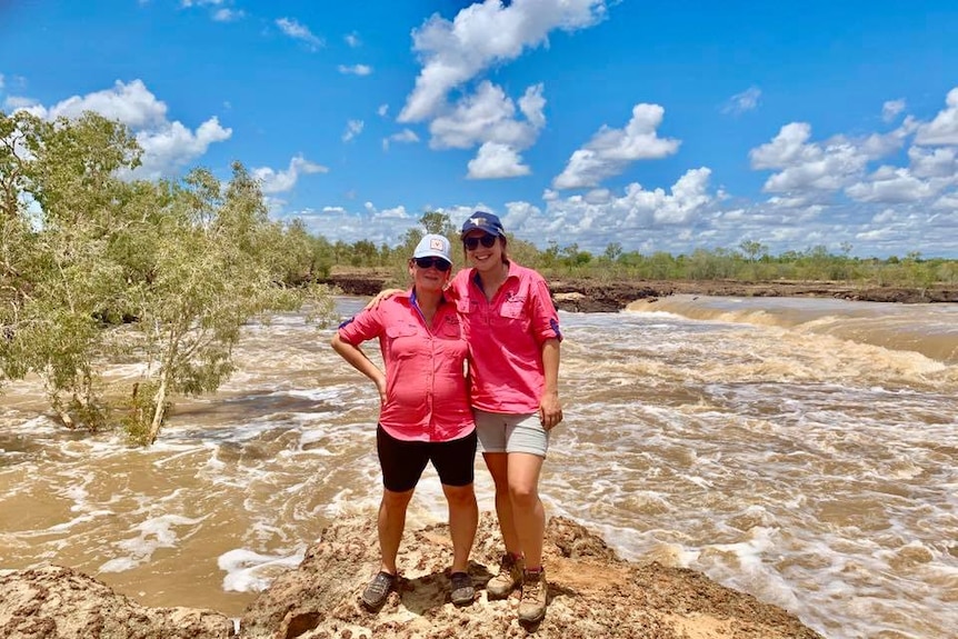 Two women dressed in pink shirts and shorts and caps stand beside a river.