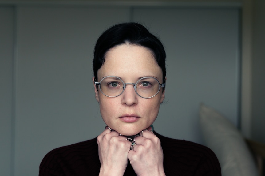 A white woman in her mid-30s with short brunette hair and glasses, hands under chin, stares at the camera 