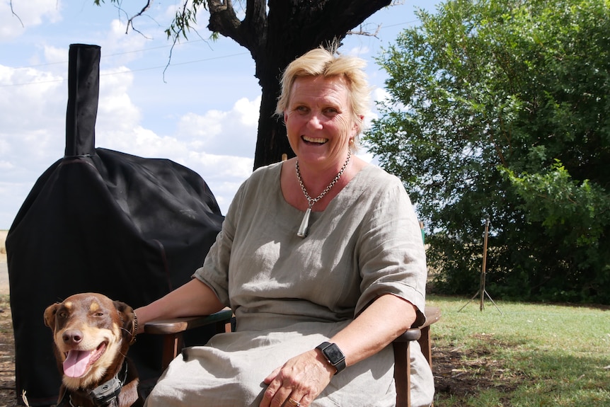 A smiling woman sits outside on a chair with a green leafy tree behind her, a brown kelpie dog sitting next to her.