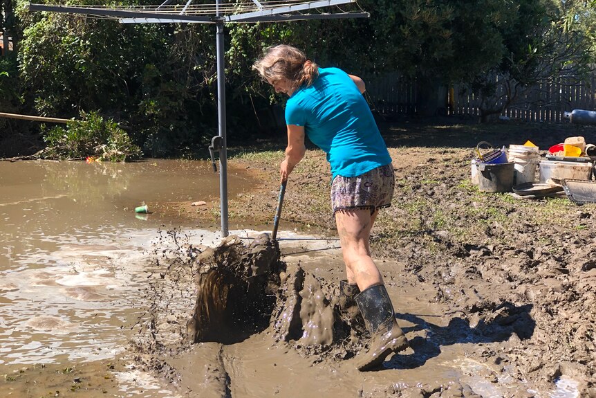 A middle-aged woman raking through mud in her flooded backyard.