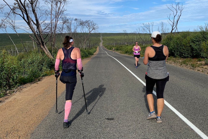 A woman with one leg and another woman walking on a bitumen road with a runner coming toward them