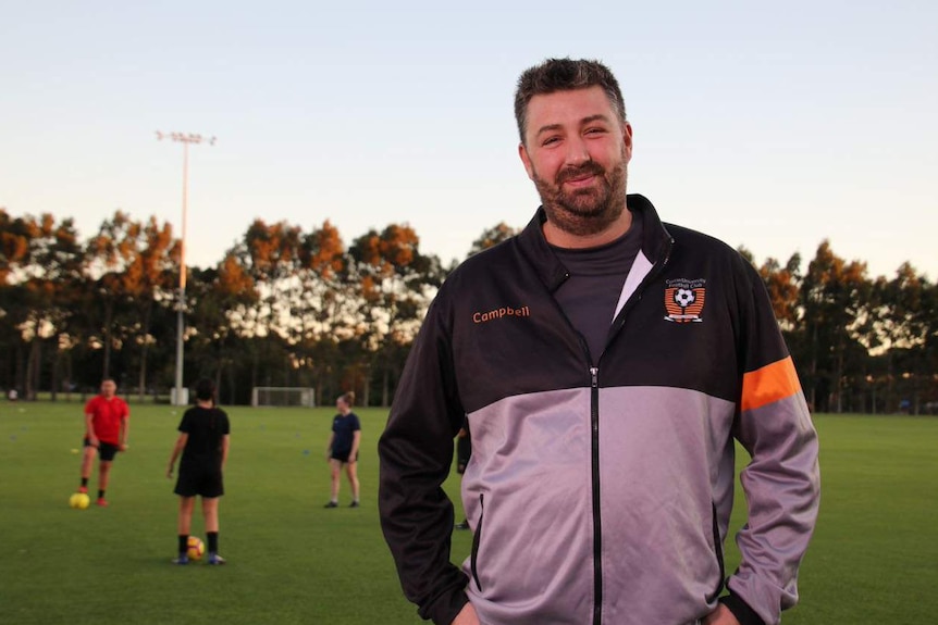 A bearded man in the left foreground in front of a sports field.