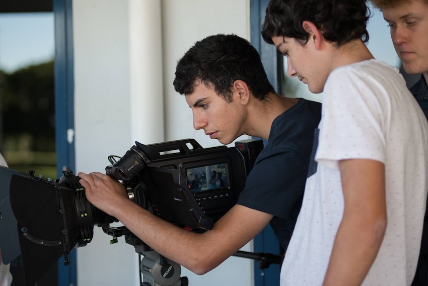 A young man adjusts focus on a movie camera lens while two colleagues look on