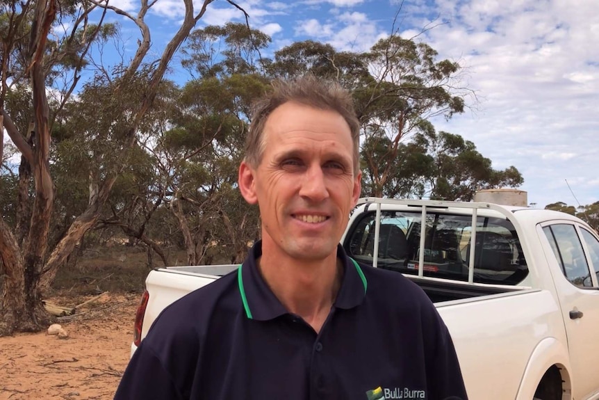 A man in a dark shirt stands in front of a ute out in the bush.