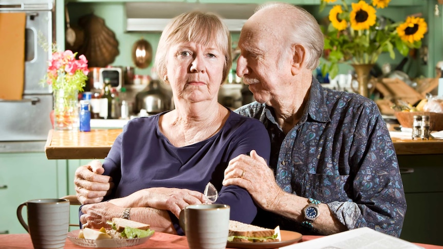 an older couple sitting at a table with sandwiches. She is looking ahead and worried, he is looking at her