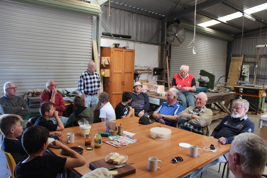 A group of boys and elderly men sit around a table with cups and tea and biscuits