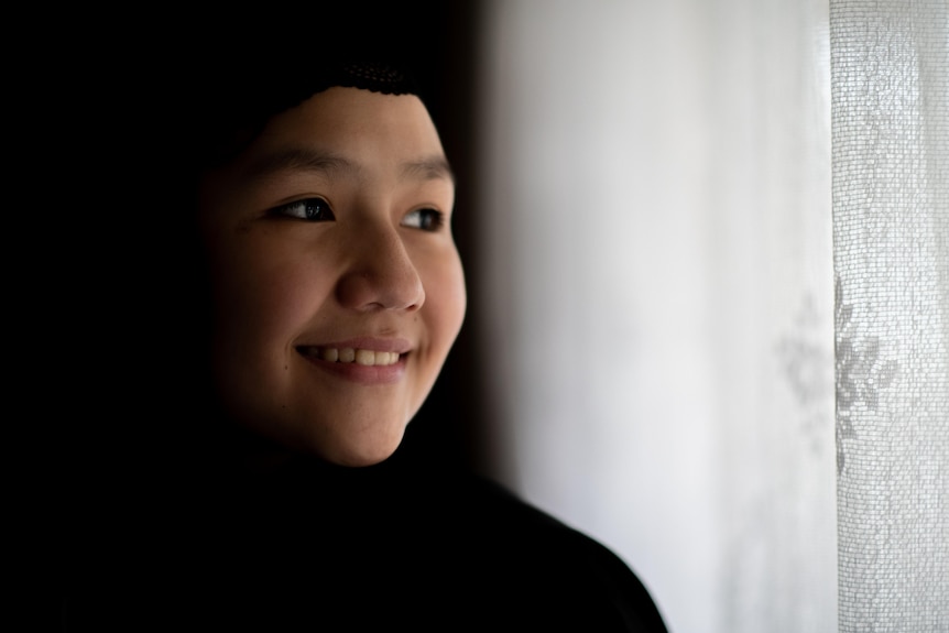 A young smiling girl stares out of a window covered with a thin white curtain