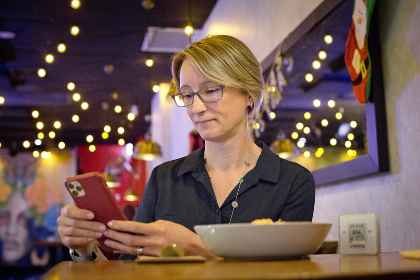 Kate Cole sits at a table inside a restaurant looking at her phone with a CO2 monitor beside her.