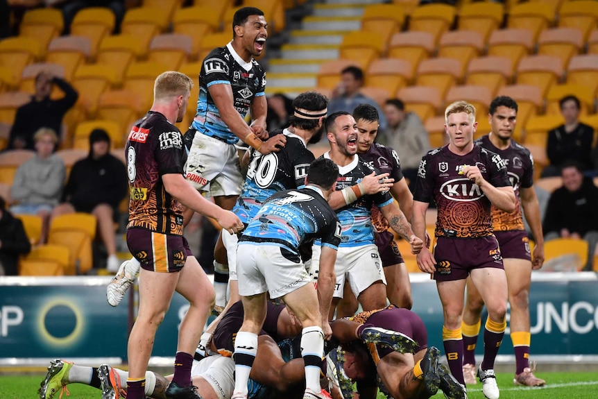 Cronulla Sharks NRL players embrace as they celebrate a try, while Brisbane Broncos opponents look on.