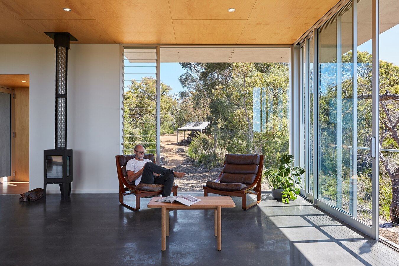 Man sits in chair house with floor to ceiling windows, light flooding in from surrounding bushland