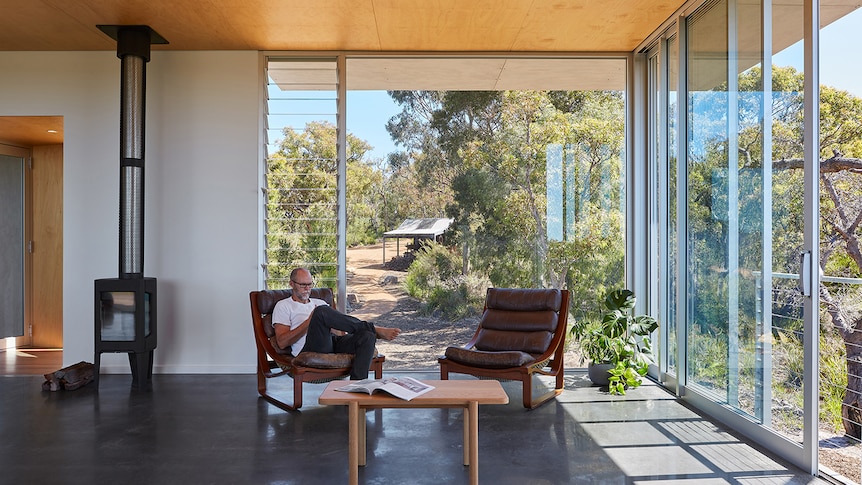 Man sits in chair house with floor to ceiling windows, light flooding in from surrounding bushland