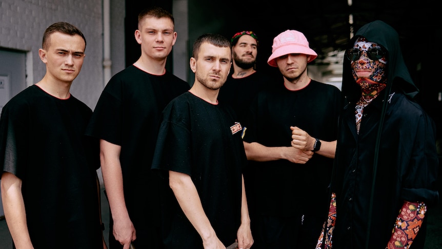 Six men standing in black shirts, one wearing a pink hat, another with tattoo sleeves on his arms and colourful mask on face