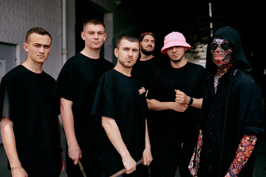 Six men standing in black shirts, one wearing a pink hat, another with tattoo sleeves on his arms and colourful mask on face
