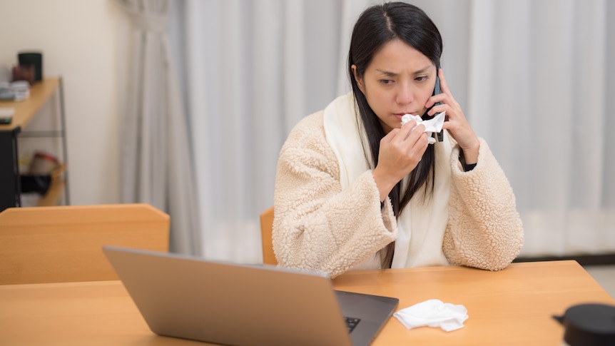 A sick-looking woman with long brown hair wipes her nose with a tissue while sitting in front of a laptop, talking on a phone.