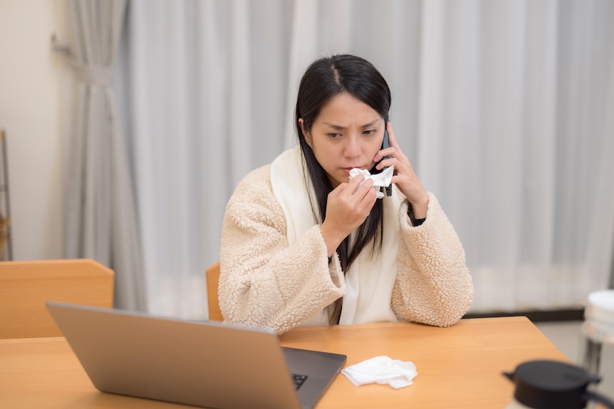 A sick-looking woman with long brown hair wipes her nose with a tissue while sitting in front of a laptop, talking on a phone.