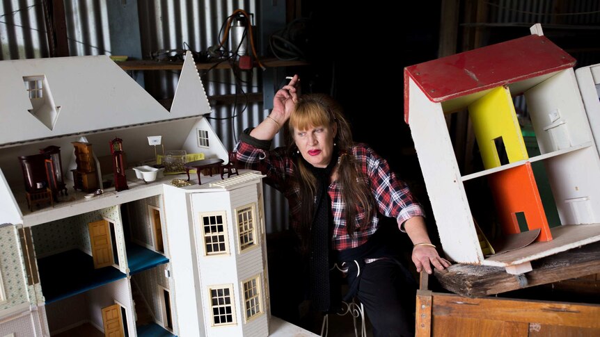 A woman leans her hand on her head while sitting among two doll houses.