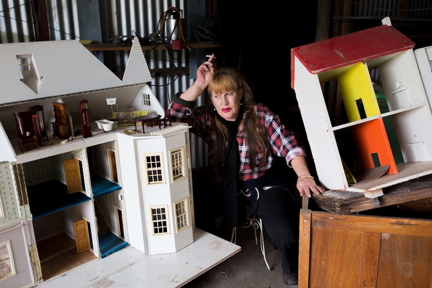 A woman leans her hand on her head while sitting among two doll houses.