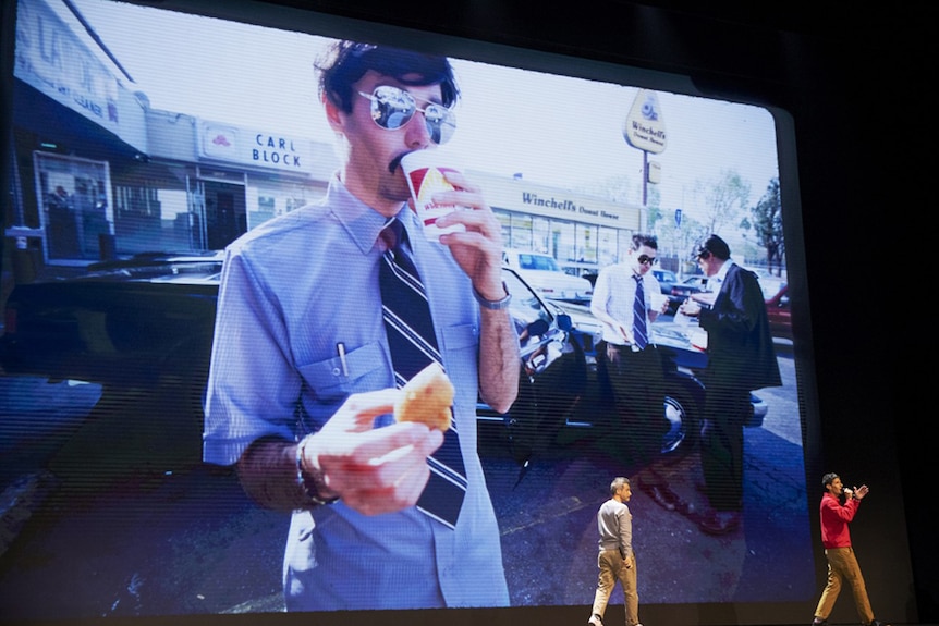 Two men on stage in front of large screen with photo of moustached man wearing sunglasses and eating and drinking in parking lot