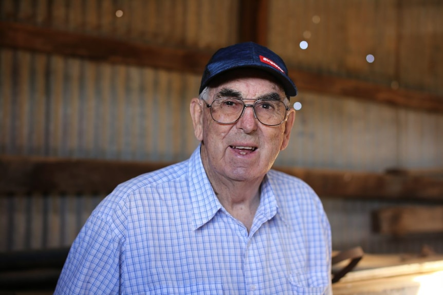 An older man in a check shirt and a cap smiles at the camera