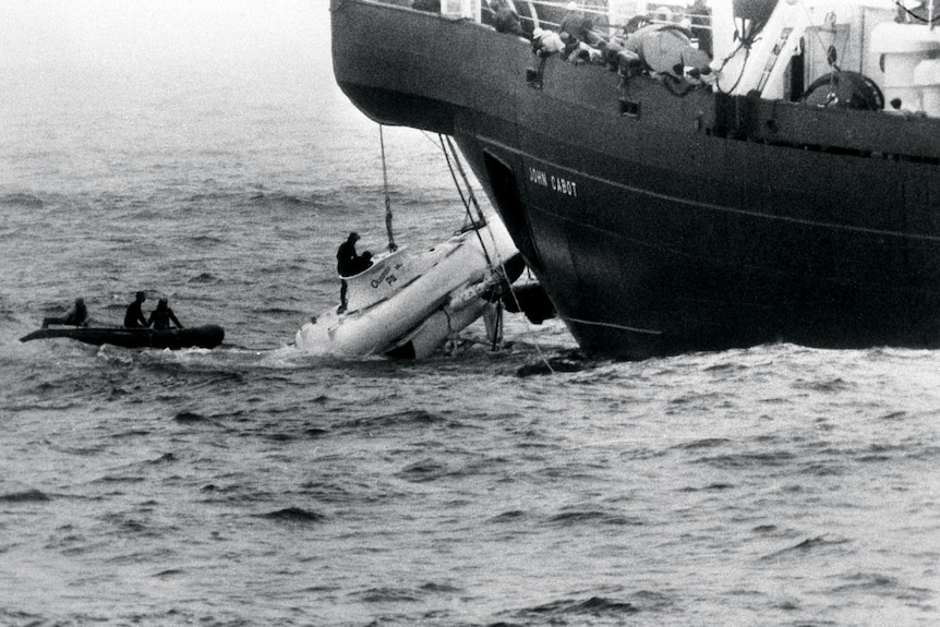 A submersible in the sea being pulled up to a large ship. People are on a small boat nearby, and a person is on the submersible