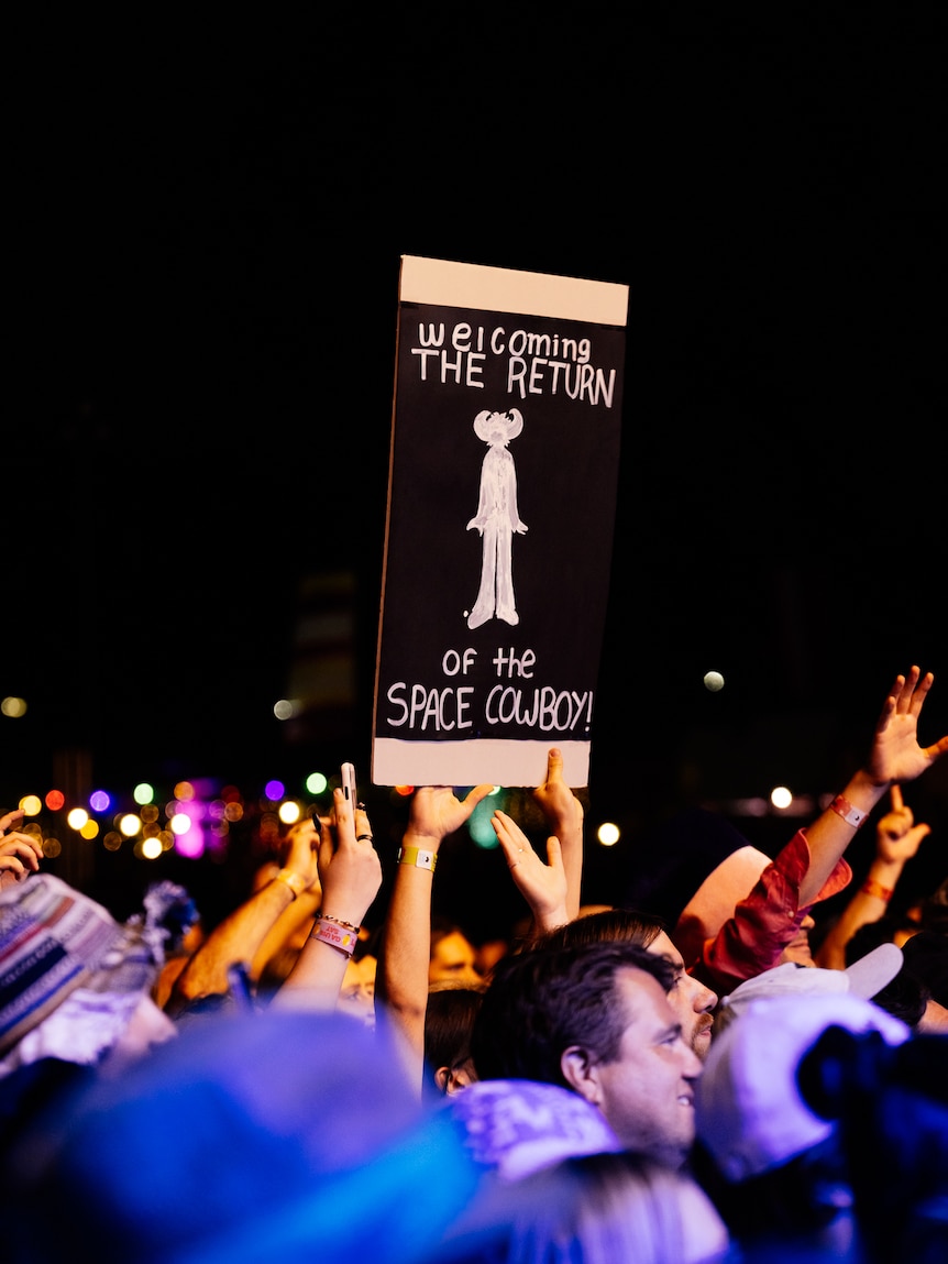 A crowd of people, someone holds up a sign saying 'Welcoming the return of the space cowboy'