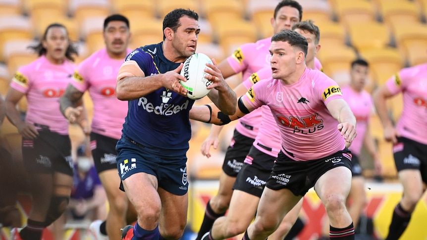 A rugby player in dark blue shirt runs the ball as opponent in pink prepares to tackle him in empty stadium.