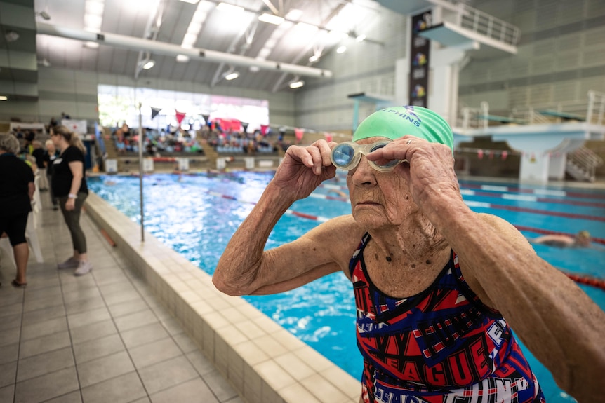 An elderly woman wearing patterned bathers and a bright green cap puts on goggles ahead of her race.