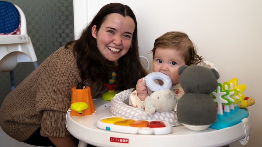 A smiling woman next to a baby in a bouncer