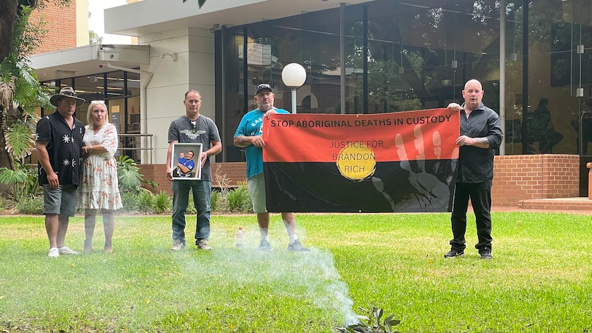 A group of people stand holding photos and a flag talking about Indigenous deaths in custody