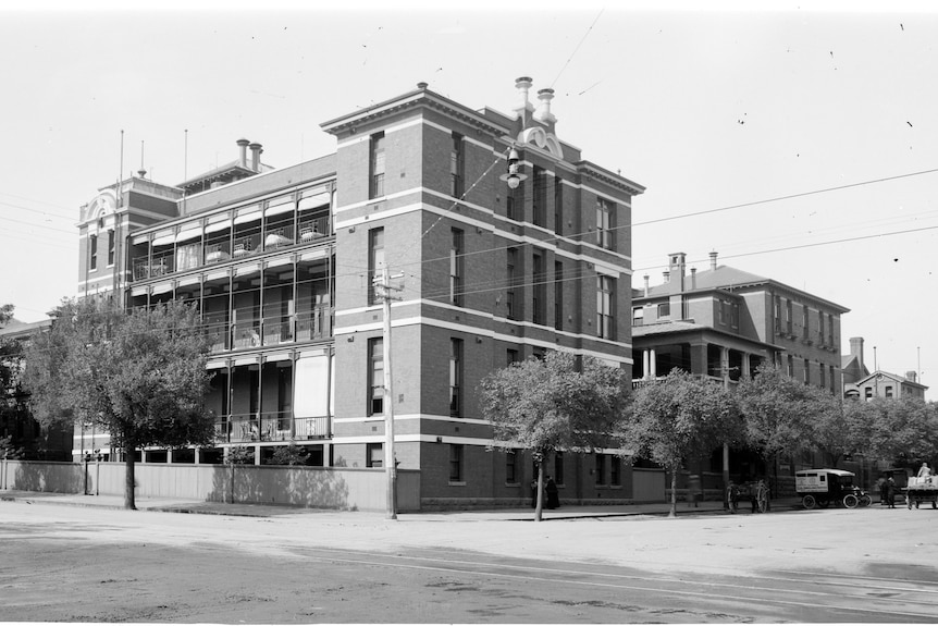 A black and white image of a three storey, brick building on a street corner.