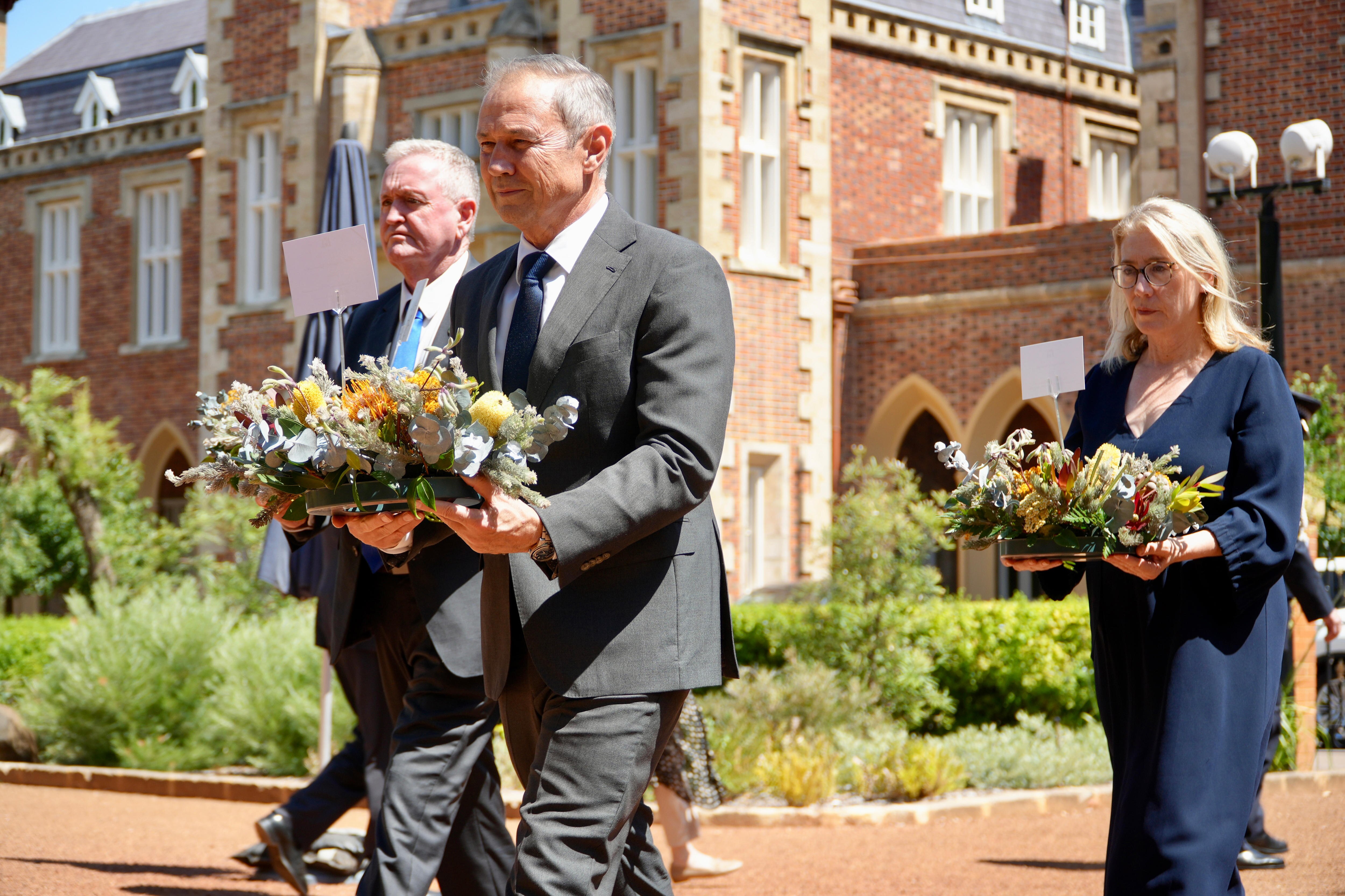 Tres personas con coronas de flores caminan frente a la Casa de Gobierno en Perth
