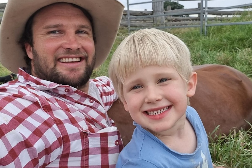 A man in a red checkered shirt smiles next to a young boy with blonde hair in a paddock