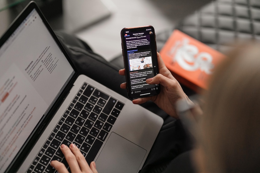 A woman holds a mobile phone in her hand while looking at a laptop.