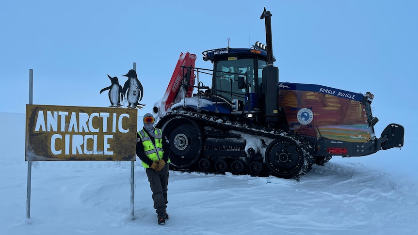 A man stands next to machinery. There is snow. He is wearing heavy snow gear and there is a sign which says Antarctica