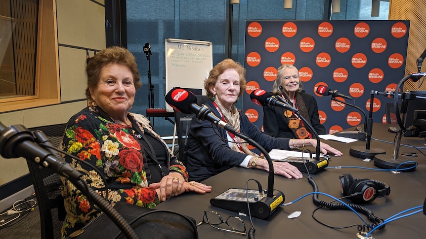 Three well-dressed elderly women sit in a row behind radio mics and smile in your direction.