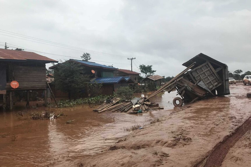 A flooded, muddy village street shows a hut that's been pushed on its side by flood waters