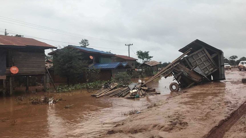 A flooded, muddy village street shows a hut that's been pushed on its side by flood waters