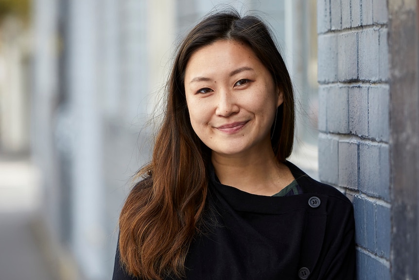 Rachel Ang smiles and wears black jumper while standing with shoulder against dark blue painted brick wall on overcast day.