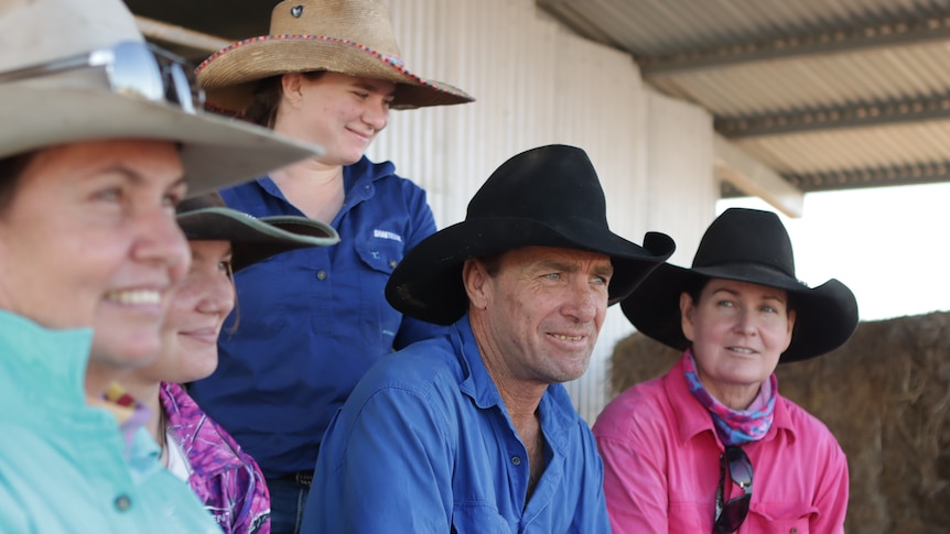 A group of people in brightly-coloured farm shirts and Akubra hats, sitting under the awning of a tin shed