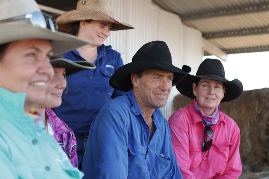 A group of people in brightly-coloured farm shirts and Akubra hats, sitting under the awning of a tin shed