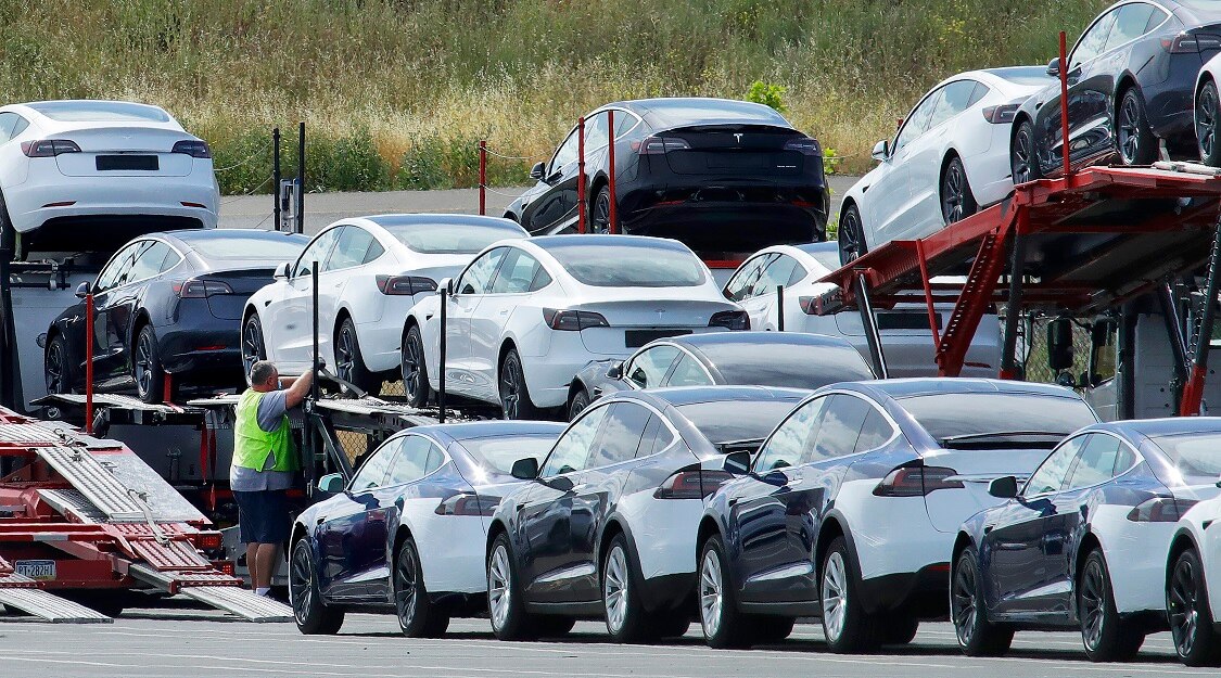 Shiny new cars are being loaded onto a transportation truck by a man in a high-visibility vest.
