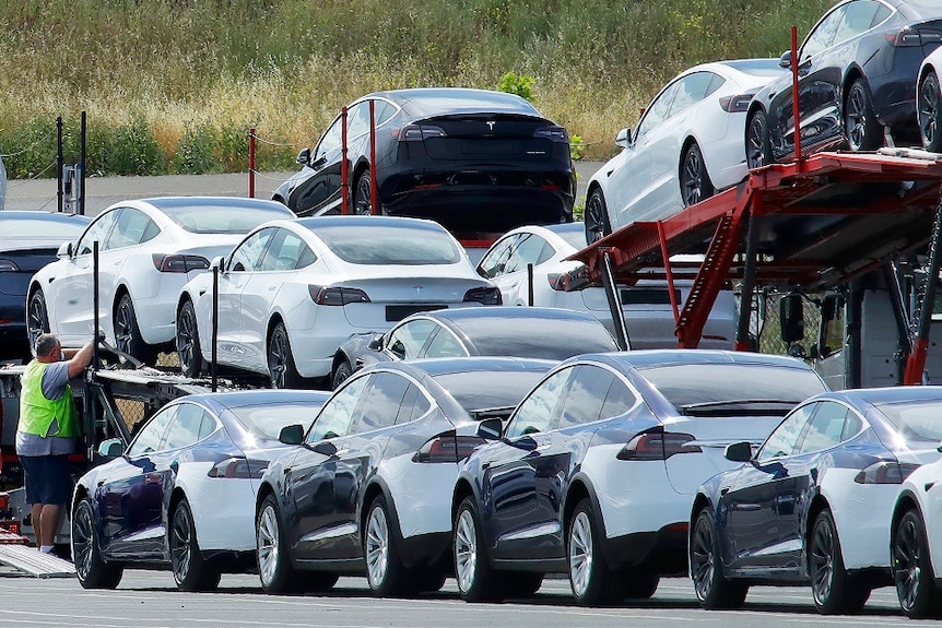 Shiny new cars are being loaded onto a transportation truck by a man in a high-visibility vest.