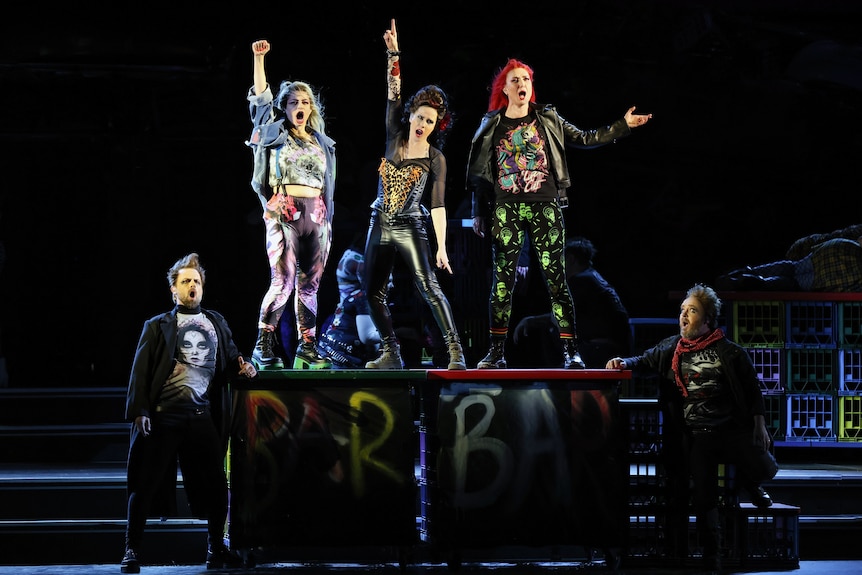Five opera singers dressed in punk streetwear perform on stage; three are on a raised platform with fists in the air.