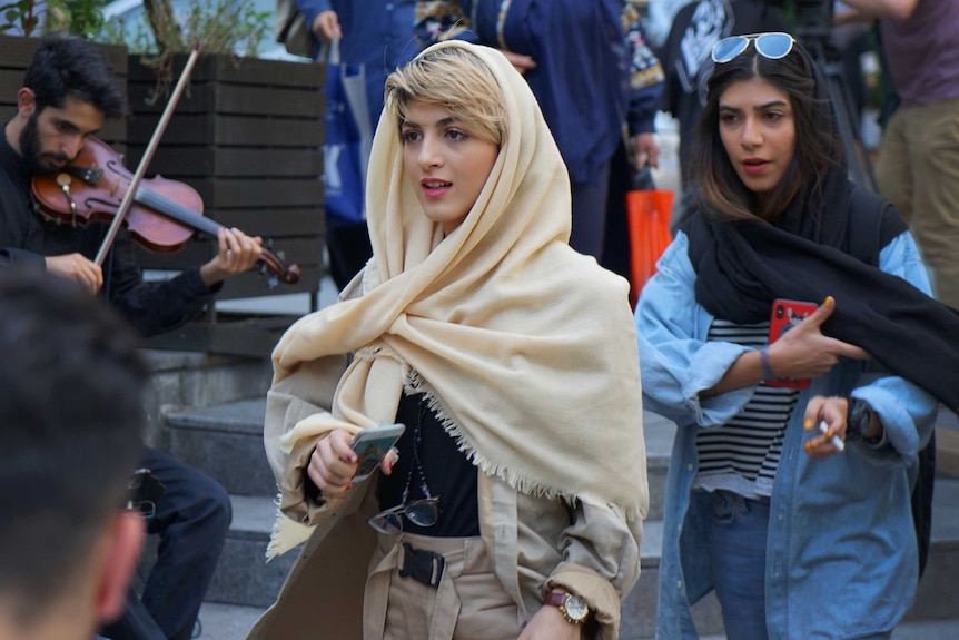 A woman with blonde hair wearing a cream scarf walks along the street.