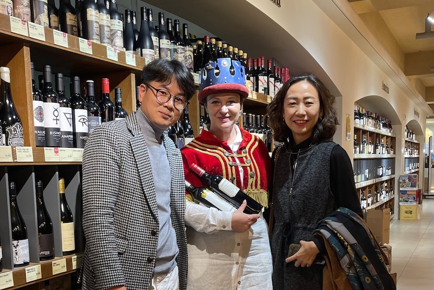 Three people standing in a bottle shop in the red wine section. Person in the middle is holding two bottles of wine. 