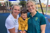 Two female Australian waterpolo players holding one of the athletes' children, smiling for a photo